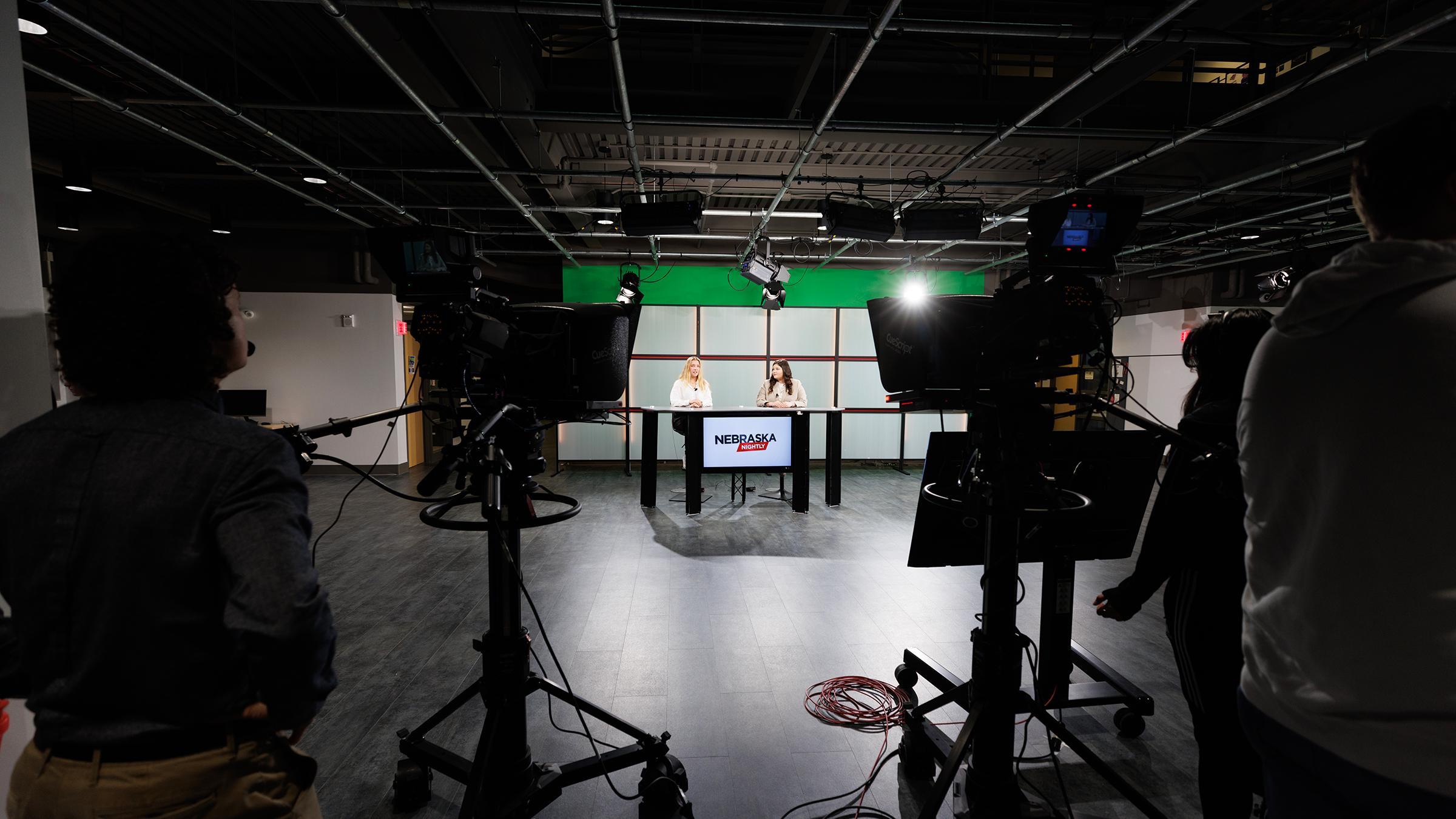 Two students sit at a news desk in a TV Studio