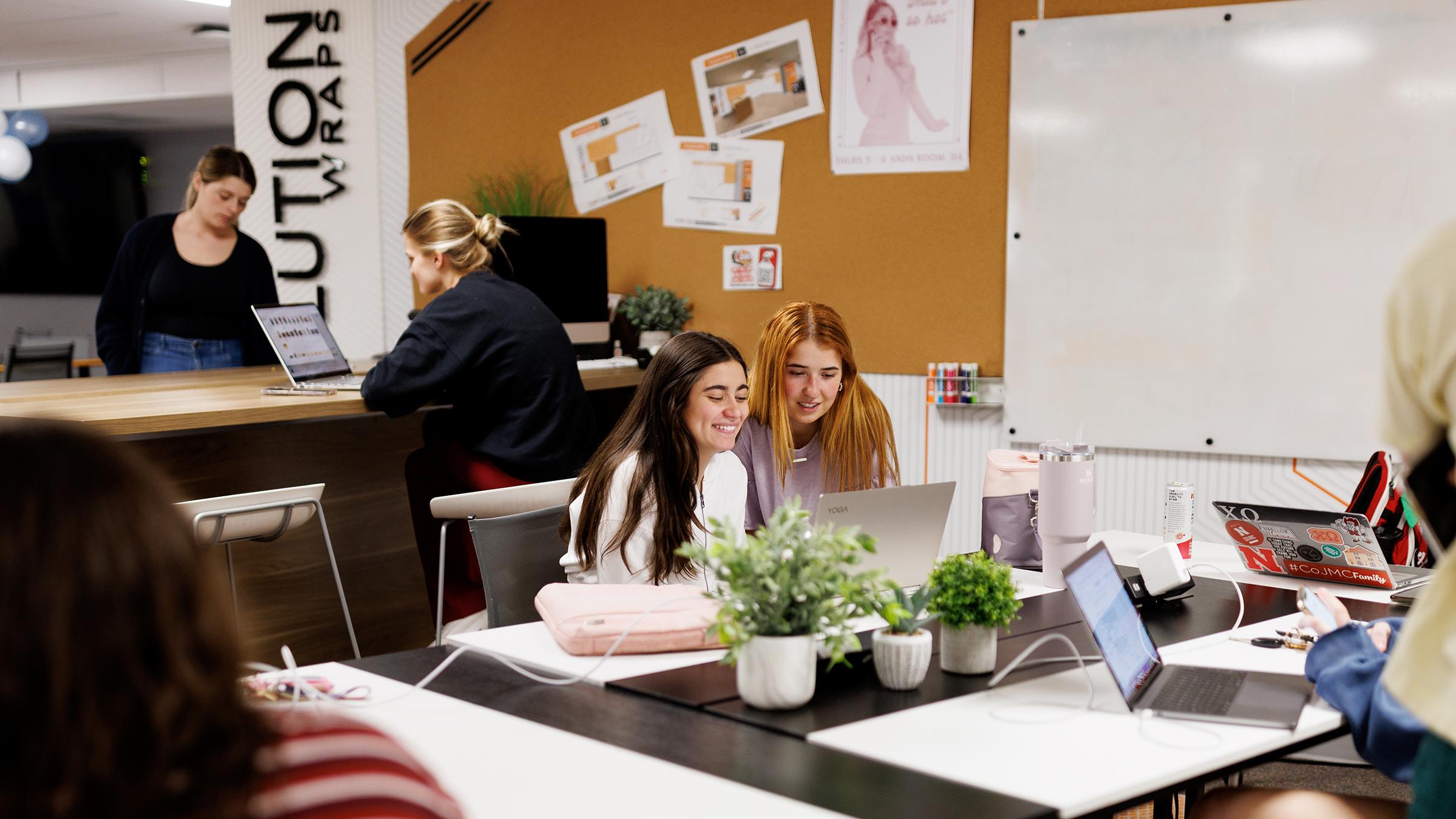 Two women look at a laptop while sitting at a desk