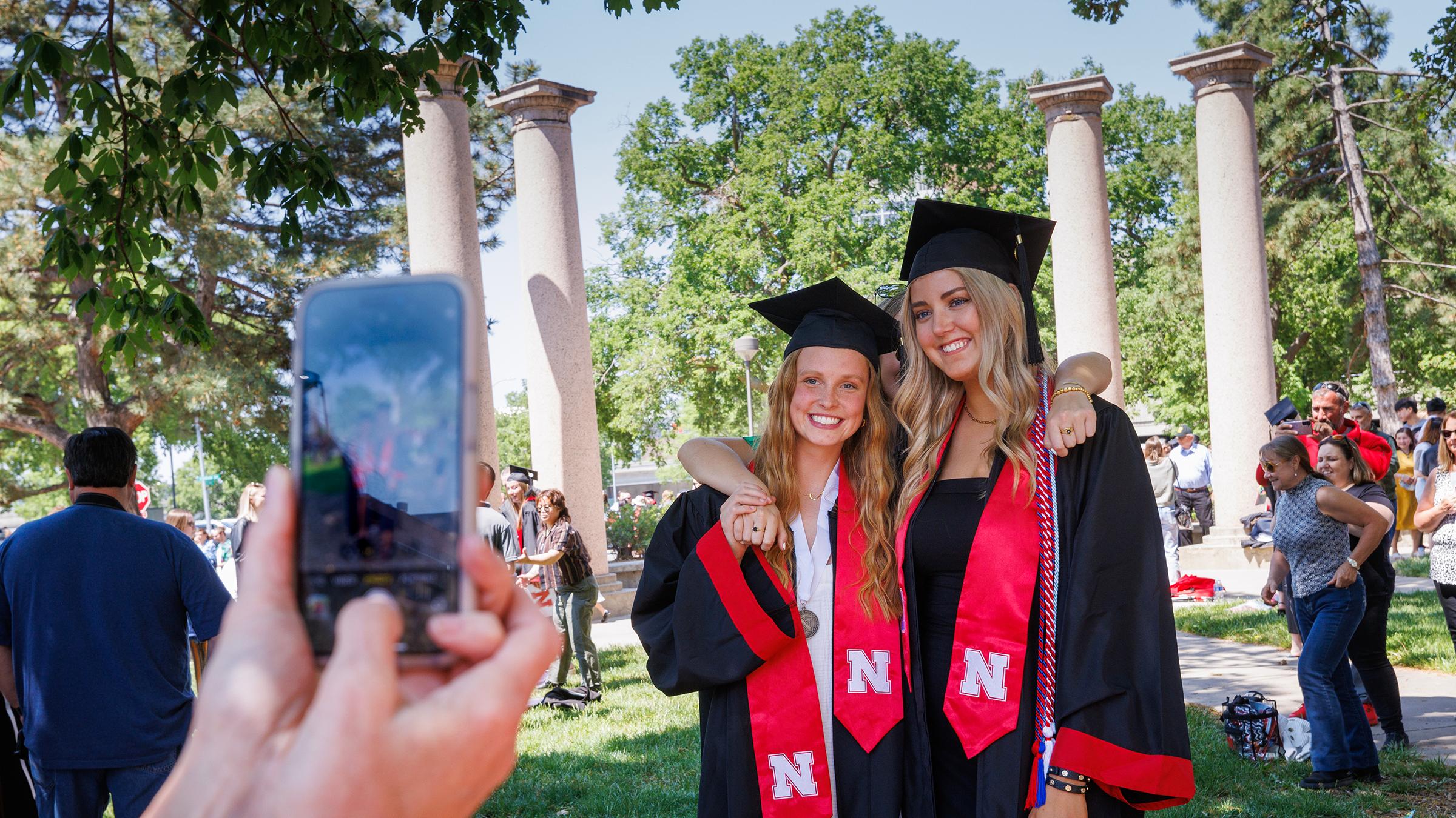 Two women pose in graduation caps and gowns while a phone takes a photograph