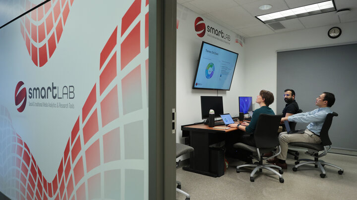 A brunette woman with short hair, brunette man with a beard and brunette man with glasses, all look up from a desk at a tv screen.