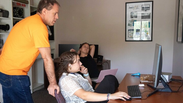 Alan Eno, associate professor of practice in broadcasting, works with Jacht crew members Hana Maddox and Molly Keifer at the Jacht Ad Agency. The three look at a computer monitor, with Eno standing and Maddox and Keifer sitting.