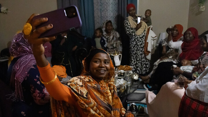 Aisha Ibrahim holds up her phone to take a selfie with a group of fellow Sudanese women and children in an apartment.