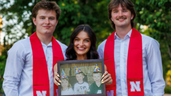 Two men in white button-ups smile next to a brunette woman holding a childhood photo of all of them in party hats.