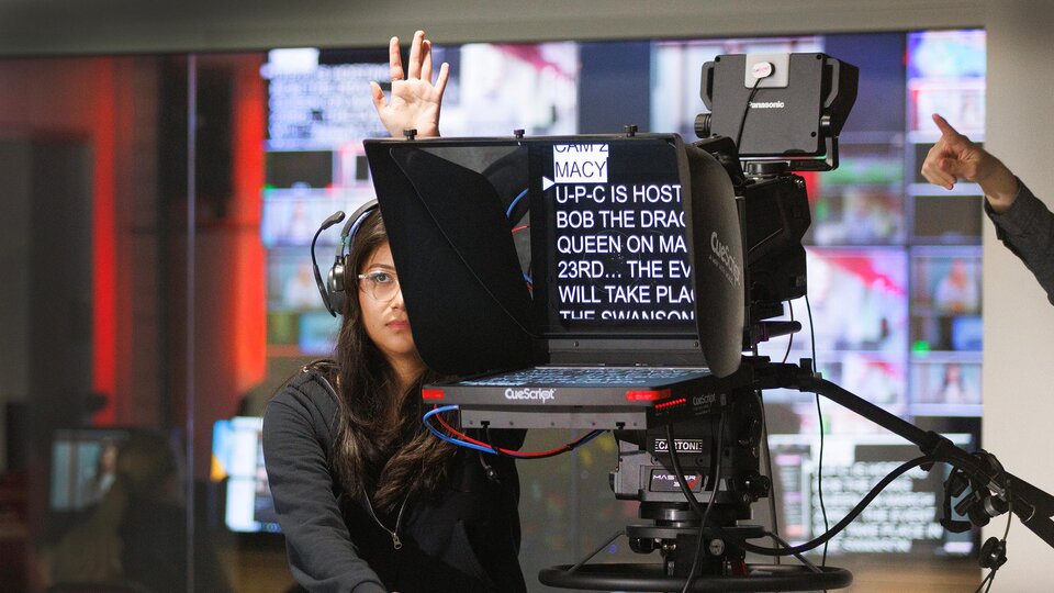 Student stands behind video camera holding up her hand