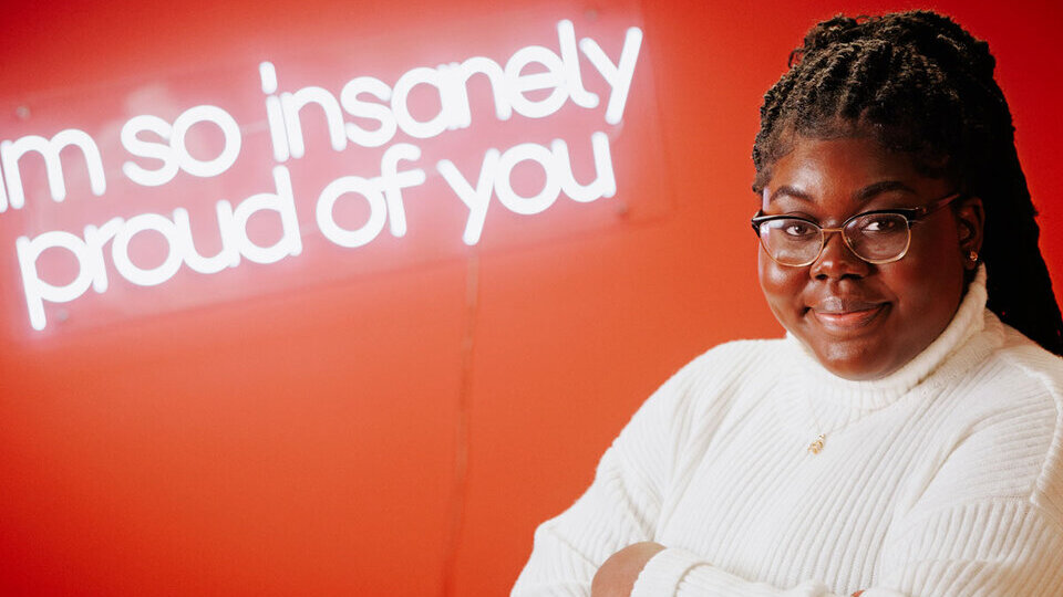 Student standing beside a neon sign that says "i'm so insanely proud of you" with her arms crossed.