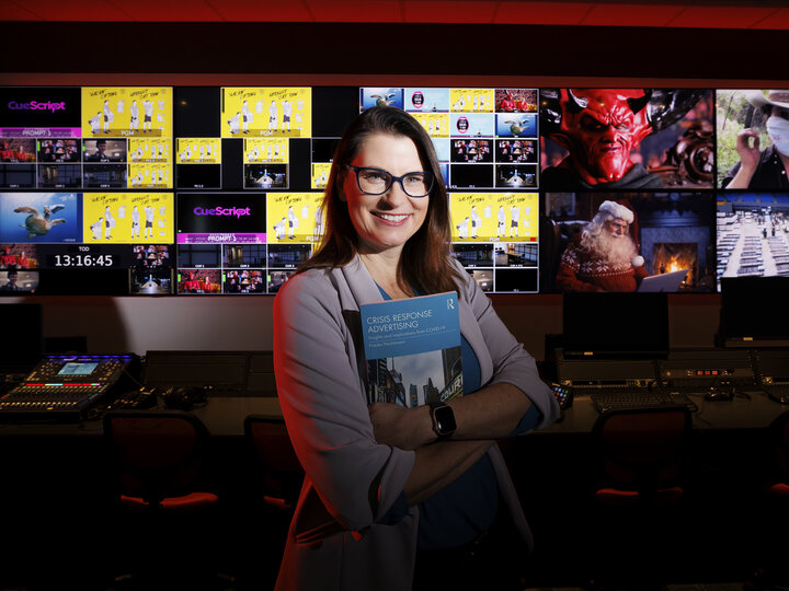 Woman stands in front of TV screens holding a book