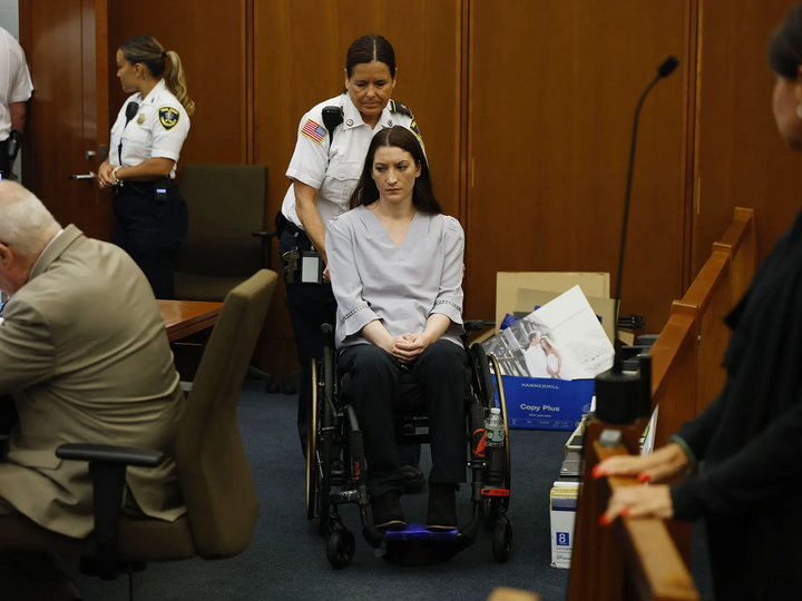 Woman in a wheelchair pushed by a guard in a courtroom setting, surrounded by people.
