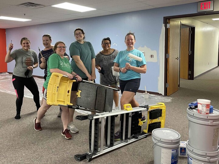 Six women hold paintbrushes and ladders while painting a classroom