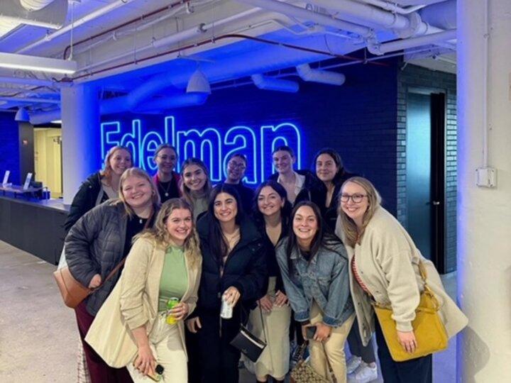 Group of students stands in front of neon sign that reads "Edelman"